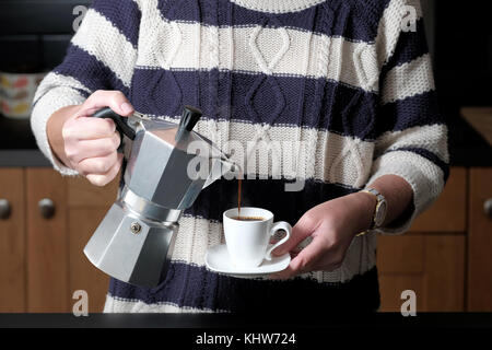 Ein smart, aber leger gekleidete Frau gießen einen Espresso aus einer molaren Herd Espressomaschine während Stehen in Ihrer Küche zu Hause. Stockfoto