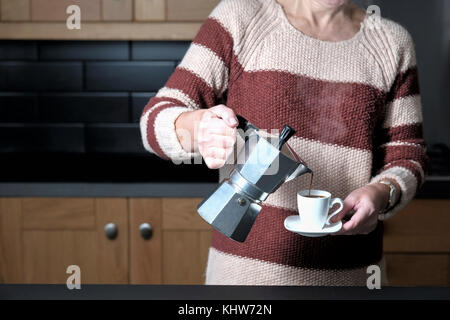 Ein smart, aber leger gekleidete Frau gießen einen Espresso aus einer molaren Herd Espressomaschine während Stehen in Ihrer Küche zu Hause. Stockfoto