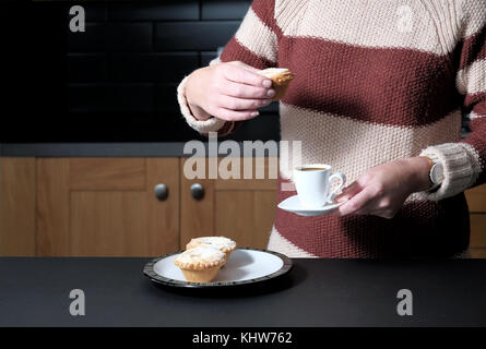 Eine kluge, aber lässig gekleidete Frau, die einen Espresso-Kaffee und einen Hackkuchen hält, während sie zu Hause in ihrer Küche steht Stockfoto