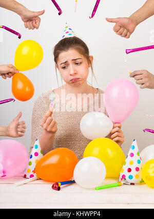 Emotionen, Gefühle, Geburtstag Konzept. sehr traurig, ungepflegtes Mädchen, schmollen, Sie durch bunte Luftballons umgeben ist, die Partei Hüte mit Punkten, Hände mit Daumen nach oben und pfeift Stockfoto