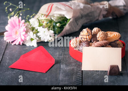 Herzlichen Glückwunsch Thema Bild mit einem leeren Papier Hinweis gegen eine Kiste voller Süßigkeiten lehnte, mit einem roten Umschlag und einen Strauß Blumen umgeben. Stockfoto