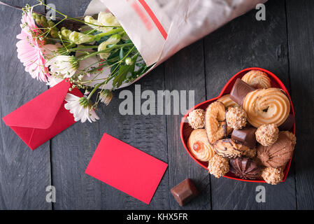 Herzlichen Glückwunsch Thema Bild mit einem roten Nachricht Karte und Umschlag, umgeben von einem Herz-förmige Kiste voller Leckereien und einen Blumenstrauß. Stockfoto