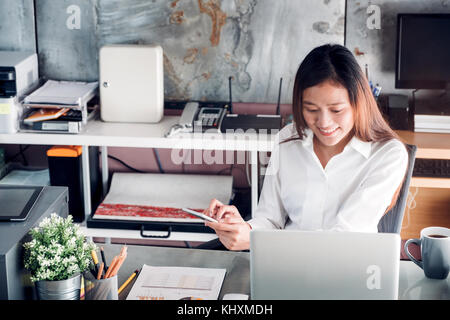 Asiatische Geschäftsfrau chattin auf Handy vor Laptop im Büro, Office lifestyle Konzept. Stockfoto