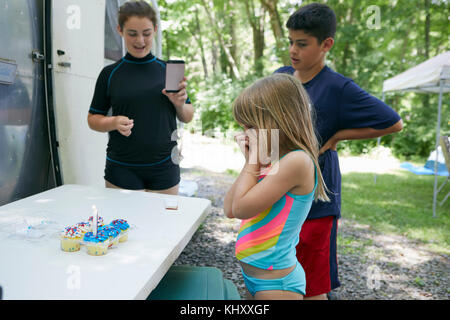 Drei Kinder standen neben Tisch mit Kuchen auf, Kerze in Kuchen, jüngere Mädchen überrascht Stockfoto