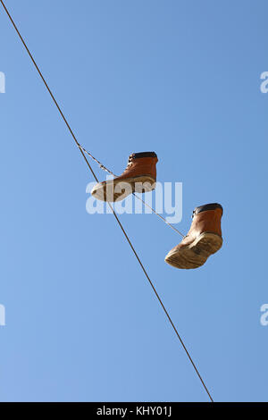 Paar alte abgenutzte braune Stiefel hängend an einer Telefonleitung Draht, wie traditionelle städtische Jugend Witz, über klaren blauen Himmel, Low Angle View Stockfoto