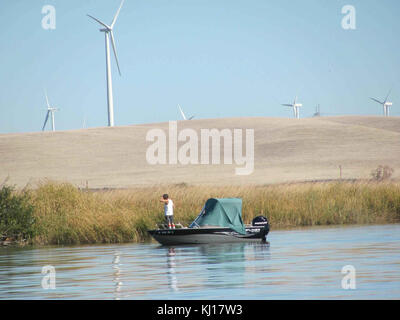 Ein Foto, das Männer in einem Boot auf einem Fluss einfängt, mit einem modernen Windpark im Hintergrund, der traditionelle und erneuerbare Energielandschaften vereint. Stockfoto