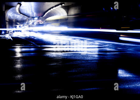 Blurry Nacht regen Verkehr auf den Straßen der Stadt. Autos in motion blur aus dem Tunnel das Licht fahren Trails und Reflexionen auf nassem Asphalt. Stockfoto