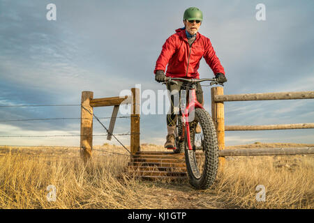 Ein älterer Mann, der auf einem Berg fat Bike über einen Cattle Guard in Soapstoneprairie natürlichen Bereich, in Northern Colorado, Ende Herbst Landschaft Stockfoto