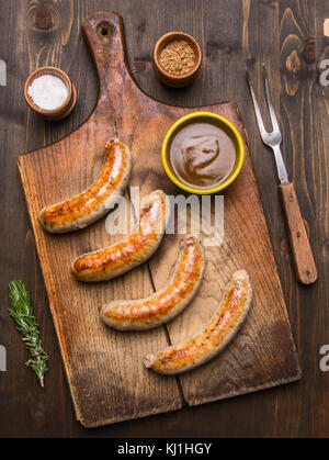 Gegrillte Würstchen liegen auf vintage Schneidbrett mit Rosmarin, Gewürzen und Fleisch Gabel auf einem urigen Hintergrund der Ansicht von oben Stockfoto