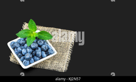 Frischen saftigen Blaubeeren auf einem dunkelgrauen Hintergrund. Heidelbeeren im weißen Quadrat Schüssel mit Minze und dekorative Matte. Stockfoto