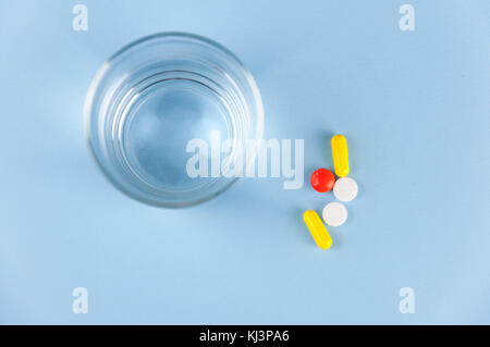 Mehrere bunte Tabletten mit Medizin liegen auf einem verschwenderischen Hintergrund, daneben ein Glas mit klarem Wasser Stockfoto