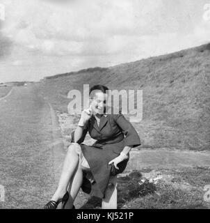 1950, historische, eine gut gekleidete junge Dame mit wohlgeformten Beine und das Tragen von high-heel Schuhe, ein smart Rock, unterrock und oben und handschuhe, sitzt neben der Straße auf einer kleinen Post für ein Bild, England, UK posieren. Stockfoto