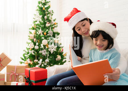 Hübsche junge asiatische Mama, ein Buch zu ihrer süßen Tochter in der Nähe von Weihnachten Baum drinnen zu Hause lesen. Frohe Weihnachten und schöne Feiertage! Stockfoto