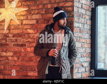 Portrait von bärtigen indischen hipster Mann in eine warme Jacke und Hut gekleidet, Getränke Kaffee aus einem roten Schale nahe der Wand eines roten Ziegeln. Stockfoto
