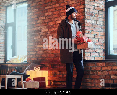 Portrait von bärtigen indischen hipster Mann in eine warme Jacke und Hut gekleidet, Getränke Kaffee aus einem roten Schale nahe der Wand eines roten Ziegeln. Stockfoto