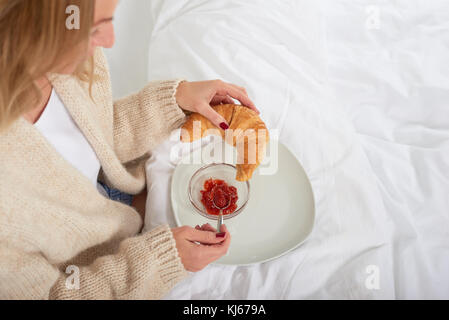 Hohe Betrachtungswinkel und einer jungen Frau essen französische Croissant mit frischem Obst, Marmelade, während Sie ein köstliches Frühstück im Bett am Wochenende Stockfoto