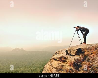 Professioneller Fotograf macht Fotos von misty Landschaft mit Spiegel Kamera und Stativ. dicker Nebel in herbstlichen Landschaft. Mann die Arbeit an scharfen clif. Stockfoto