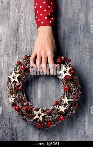 Nahaufnahme eines jungen kaukasischen Frau trägt ein rotes Kleid mit weißen Punkten Holding a christmas Kranz in der Hand auf einem grauen Holzmöbeln im Landhausstil suchspray Stockfoto
