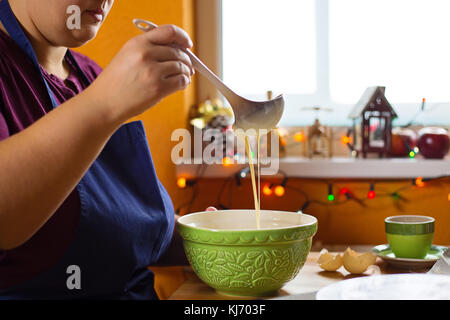 Nahaufnahme Foto der jungen Frau sitzt in der Küche hinter einem Tisch mit einer Schüssel voll mit Flüssigkeit Pfannkuchen Teig. Sie prüft die Dichte der Teig Stockfoto