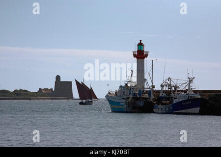 Hafen von St Vaast: Leuchtturm, moderne Fischerboote, die mit Anker liegen und traditionelle Bisquine 'Ami Pierre', die den Hafen unter Segeln verlassen, Normandie, Frankreich Stockfoto