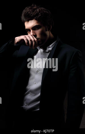 Denken charismatische Mann posiert und Suchen mit der Hand in der Nähe der Fläche auf dunklen Schatten dramatischen Licht Hintergrund. Closeup Portrait. Stockfoto