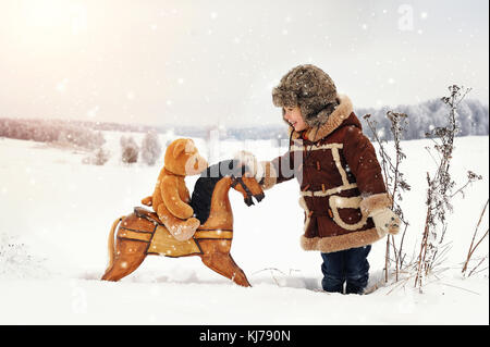 Ein lächelndes, vier Jahre alten Jungen tragen Schaffellmantel spielen mit Holz Schaukelpferd in schneebedeckten Feld. Stockfoto