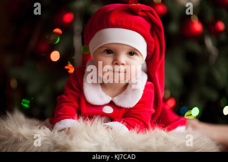 Fröhliches Baby Mädchen in santa claus Kostüm und Hut liegt auf Fell Teppich gegen den Hintergrund von Weihnachten Dekorationen und Lichter. closeup. Stockfoto