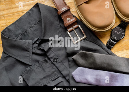 Männer Kleidung auf einem Holz- Oberfläche, zeigt ein schwarzes Kleid Shirt, braun Ledergürtel, braun Stiefel, Krawatten und Schwarze Armbanduhr Stockfoto