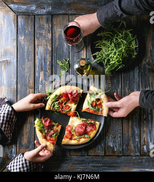 Hände, die in Scheiben geschnittene hausgemachte Pizza mit Käse und Bresaola, auf schwarze Platte serviert mit frischem Rucola, Olivenöl, Rotwein und Küchenpapier Stockfoto