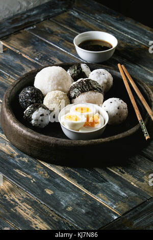 Stein Fach mit unterschiedlicher Größe Reisbällchen mit schwarzem Sesam und Algen Nori, serviert mit weich gekochte Eier, Sojasauce, Essstäbchen auf Holzbrett ba Stockfoto