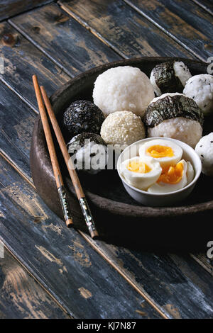 Stein Fach mit unterschiedlicher Größe Reisbällchen mit schwarzem Sesam und Algen Nori, serviert mit weich gekochte Eier und Essstäbchen auf Holzbrett Hintergrund Stockfoto