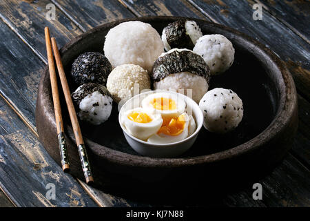 Stein Fach mit unterschiedlicher Größe Reisbällchen mit schwarzem Sesam und Algen Nori, serviert mit weich gekochte Eier und Essstäbchen auf Holzbrett Hintergrund Stockfoto