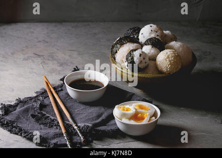 Schüssel mit Reis Kugeln verschiedener Größe mit schwarzem Sesam und Algen Nori, serviert mit weich gekochte Eier, Sojasauce, Stäbchen über graue Tabelle. Asiatische sty Stockfoto