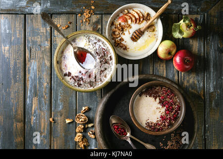 Vielzahl Schüsseln Milch Getreide Brei mit verschiedenen Zusatzstoffen, serviert mit Äpfeln, Beeren und Samen über alte hölzerne Planke Hintergrund. Ansicht von oben mit der Stockfoto