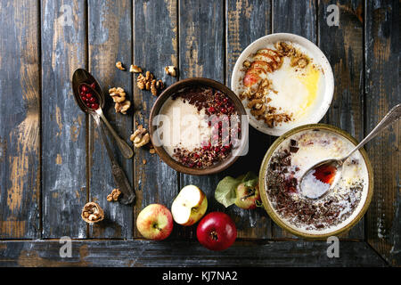 Vielzahl Schüsseln Milch Getreide Brei mit verschiedenen Zusatzstoffen, serviert mit Äpfeln, Beeren und Samen über alte hölzerne Planke Hintergrund. Ansicht von oben mit der Stockfoto