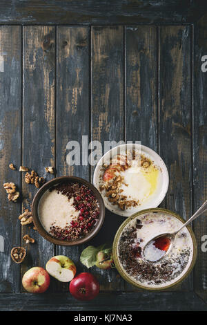 Vielzahl Schüsseln Milch Getreide Brei mit verschiedenen Zusatzstoffen, serviert mit Äpfeln, Beeren und Samen über alte hölzerne Planke Hintergrund. Ansicht von oben mit der Stockfoto