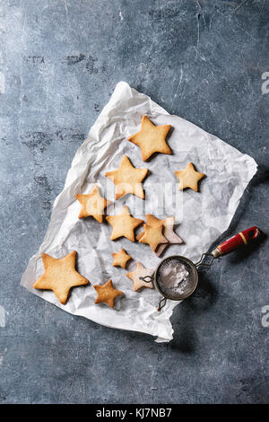 Hausgemachte Kekse Stern Sugar Cookies unterschiedlicher Größe mit Zucker Pulver auf Backpapier mit Vintage Sieb über blaue Textur Hintergrund. Christm Stockfoto