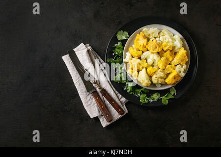 Weiß-gelbes Blumenkohlgratin auf schwarzem Keramikteller mit Korianderblättern, textiler Serviette und Besteck auf dunklem Texturhintergrund. Flach Stockfoto