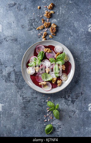 Vegan rote Beete carpaccio Salat mit Radieschen, Basilikum, Olivenöl und Nüssen in Keramik Platte über blaue Textur Hintergrund. Ansicht von oben, Platz. Gesunde eatin Stockfoto