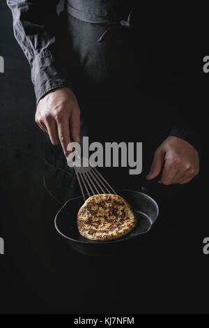 Mann Koch in schwarze Schürze kochen Pfannkuchen in gusseisernen Pfanne. Schaltet die Pfannkuchen. Dunklen rustikalen Stil. Stockfoto