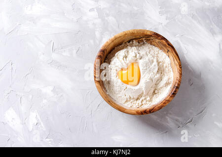 Olivenholz Schüssel mit Weizenmehl und ganze Eigelb als Herzform über grau Textur Hintergrund. Ansicht von oben mit der Kopie. Backen Konzept Stockfoto