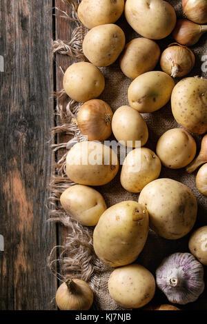 Rohe ganze gewaschen Bio Kartoffeln, Zwiebeln und Knoblauch in Sacktuch über alte Holzbrett Hintergrund. Ansicht von oben mit dem Raum. Nahaufnahme Stockfoto