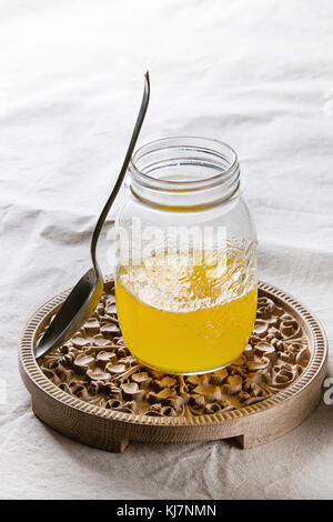 Glas heisses gezeichnet ghee Butter stehend mit Löffel auf Holz verzierten Brett über weiße Bettwäsche textil Tischdecke. Gesunde Ernährung Stockfoto