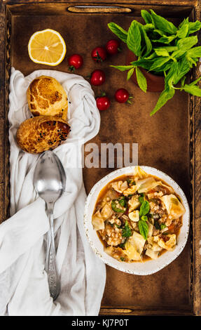 Kohlsuppe in einer weißen Schüssel in eine hölzerne Fach von oben fotografiert. Frische Minze, Abendessen Brötchen, Tomaten, Zitrone und ein Löffel o Stockfoto