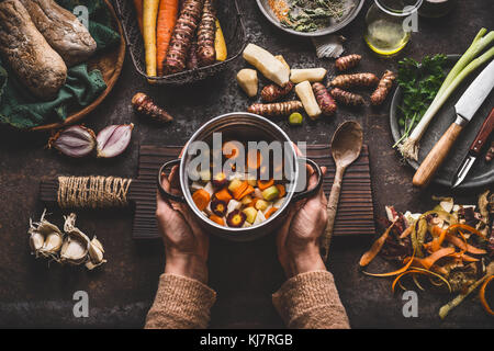 Weiblich frau Hände halten Pan mit gewürfelten bunte Gemüse auf dunklen rustikalen Küche Tisch mit vegetarische Küche Zutaten und Werkzeuge. gesund und Stockfoto