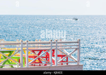 Bunte rot, grün und gelb lackierten Tische auf der Veranda, Terrasse und Balkon von Heim, Haus mit Blick auf den Ocean View mit Boot und Zaun Geländer im Sommer r Stockfoto