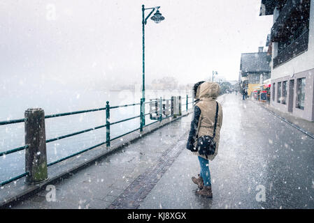 Winter Wetter Thema Bild mit einer Frau gekleidet für kalte, Wandern auf der Straße, unter dem Schneefall, in der Nähe des Hallstätter See und Hallstatt Stadt, aus Stockfoto