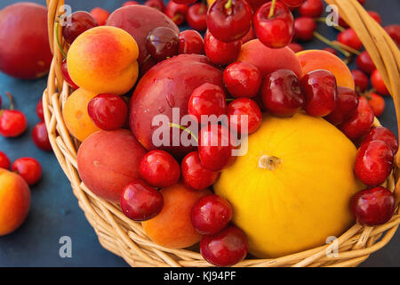 Reif organische Obst Kirschen Nektarinen Aprikosen lebendigen Farben in Weidenkorb auf dunklem Blau Tischplatte verstreut Melone. Sommer Ernte sauber Essen Stockfoto