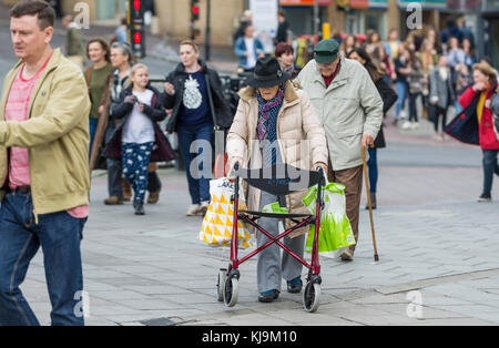 Ältere Dame zu Fuß durch einen Beschäftigten überfüllten Stadt mit Hilfe einer fahrbaren Gehhilfe in England, Großbritannien. Ältere Frau mit einem Rollator Gehhilfe. Stockfoto