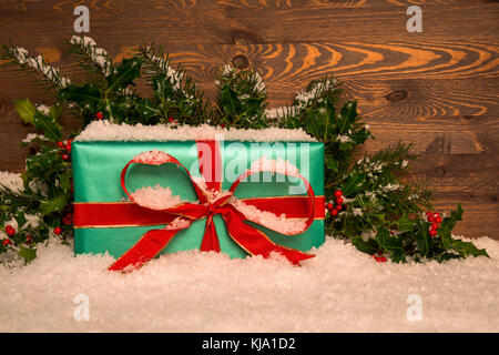 Weihnachtsgeschenk Geschenk im Grünbuch mit roter Schleife verpackt mit Stechpalme und Schnee gegen eine Holz- Hintergrund, kopieren Sie Platz für Ihre Nachricht. Stockfoto